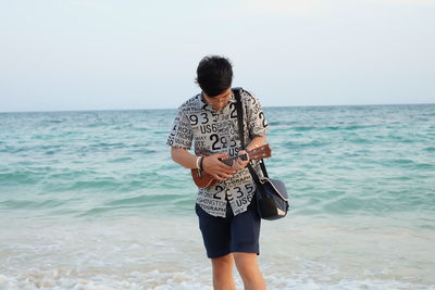 Young man holding violin while standing at beach against sky during sunset