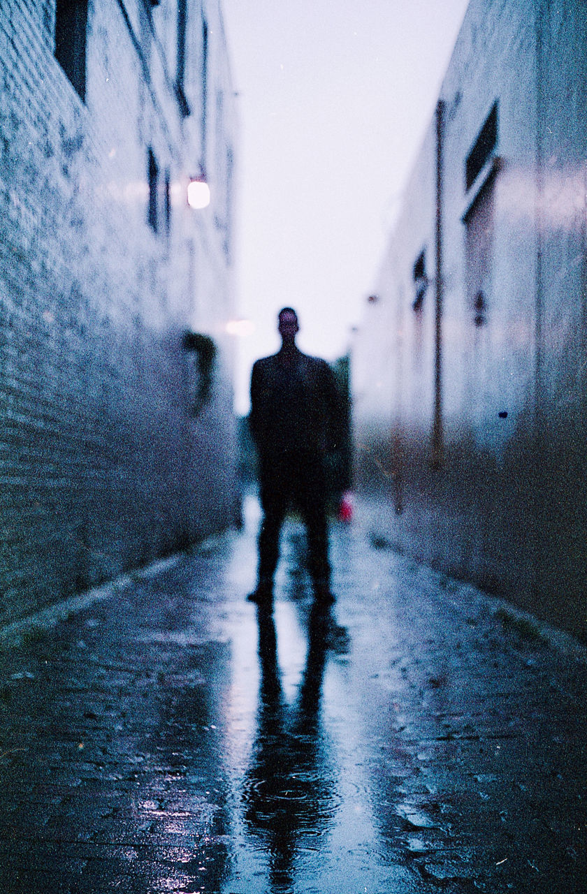 REAR VIEW OF MAN WALKING ON STREET AMIDST BUILDINGS