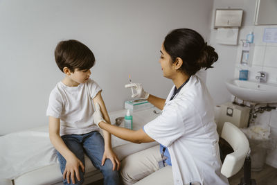 Side view of female pediatrician giving vaccine to boy sitting on bed in clinic at hospital