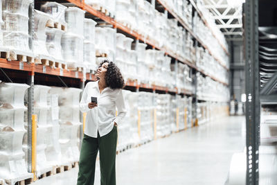 Businesswoman with smart phone standing by rack in warehouse