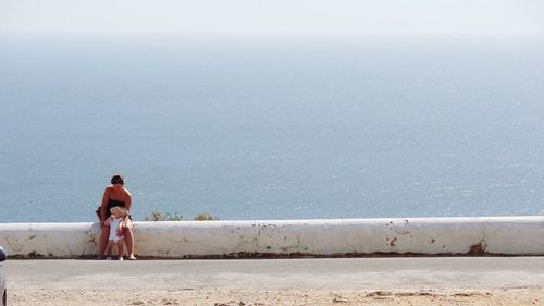 Mother and child sitting on retaining wall against sea