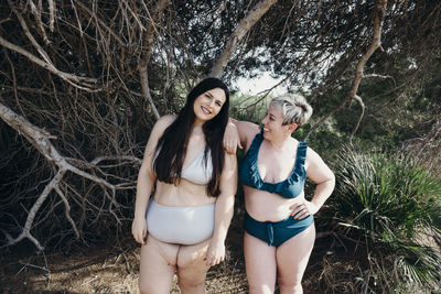 Cheerful curvy female friends in swimwear standing looking away on beach near trees in summer