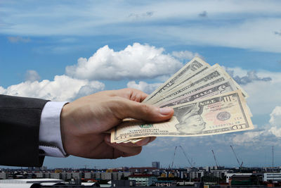 Cropped hand businessman holding paper currency against sky