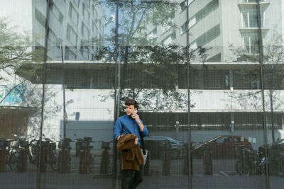 Young businessman leaning on wall of modern glass building while talking on smart phone