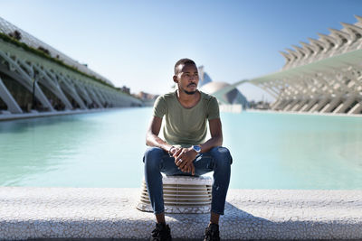 Thoughtful african american male in casual wear relaxing near fountain in city on sunny day and looking away