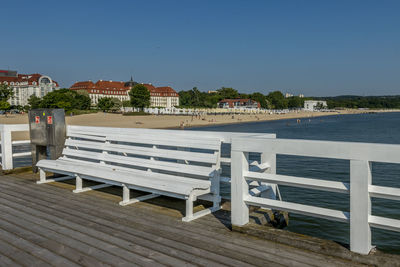 Bench by sea against buildings against clear blue sky