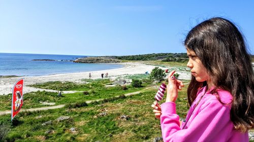 Girl holding ice cream while looking away at beach against clear blue sky