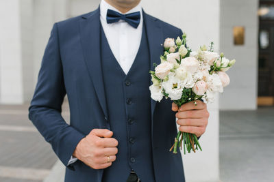 The groom in a blue suit holds in his hand a delicate wedding bouquet for his beloved bride