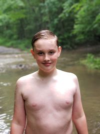 Portrait of smiling shirtless boy standing against stream in forest