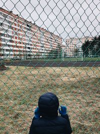 Rear view of man looking through chainlink fence