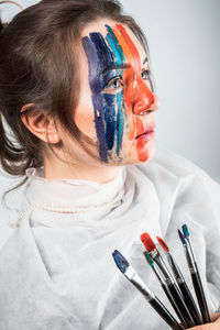 Young woman with painted face holding paintbrushes against gray background