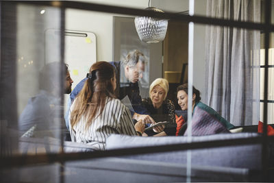 Businessman showing digital tablet to colleagues in board room during meeting seen from glass