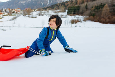 Child in blue clothes climbing up the snowy hill pulling the red sled