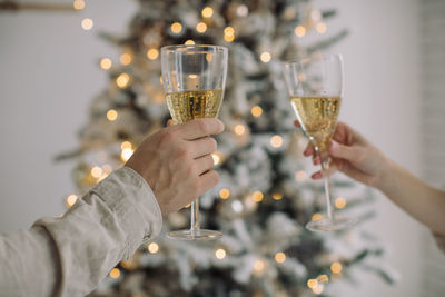 Cropped hand of couple holding champagne flutes against christmas tree at home