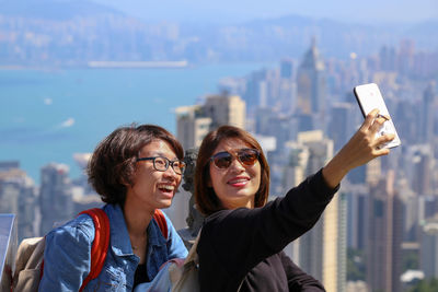 Smiling friends taking selfie while standing against modern cityscape