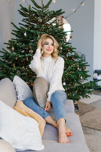 Lovey woman is sitting on a gray sofa near the christmas tree in the living room and holding 