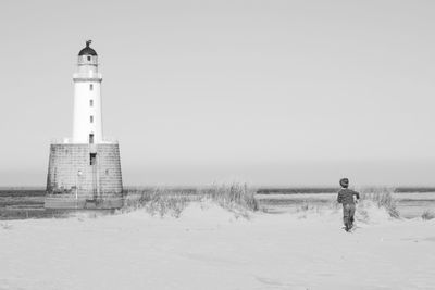 Child runs towards lighthouse by sea against clear sky