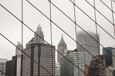 Low angle view of buildings against sky behind brooklyn bridge cables