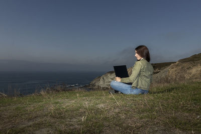 Young woman, the freelancer sitting on a high cliff over the sea and works remotely