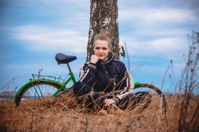 Portrait of woman with bicycle sitting by tree trunk against sky