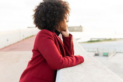 Side view of african american female with afro hairstyle wearing jacket leaning on barrier of embankment of river and looking away