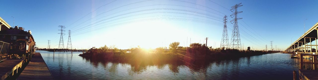 water, sun, clear sky, reflection, sunlight, built structure, sunbeam, waterfront, sunset, building exterior, power line, electricity pylon, transportation, blue, sky, river, architecture, lens flare, lake, nature