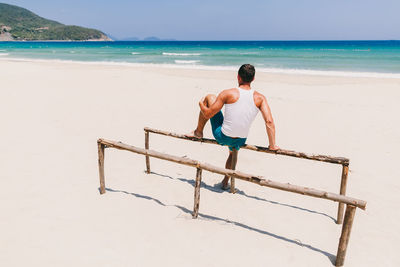 Rear view of man sitting on parallel bars at beach during sunny day