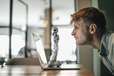 Man looking at toy robot, standing on his laptop