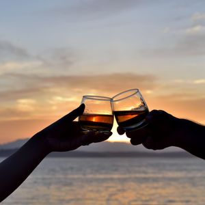 Cropped silhouette hands of couple toasting drinks at beach against sky during sunset