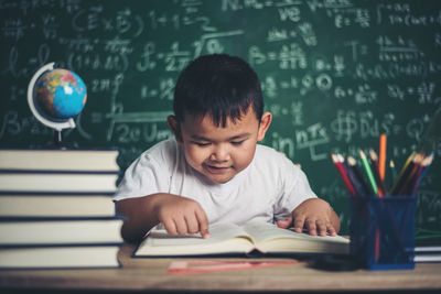 Cute boy studying at table against blackboard