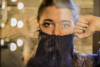 Portrait of woman covering face with turtleneck sweater seen through glass