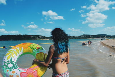 Rear view of young woman in bikini carrying inflatable ring at beach