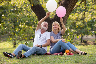 Smiling couple holding balloons while sitting by fruits on grass