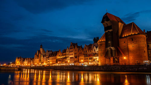 Illuminated buildings by river against sky at dusk