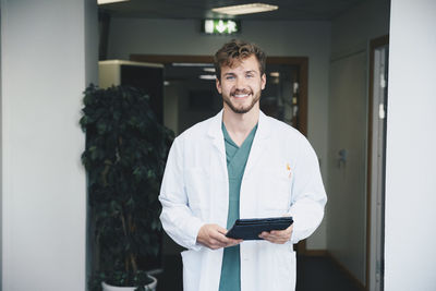 Portrait of confident smiling male doctor standing with digital tablet at hospital corridor