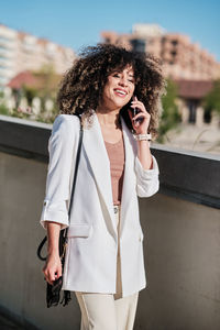 Merry hispanic female entrepreneur in white suit with curly hair smiling and talking on smartphone while walking near fence on sunny day on city street