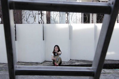 Young woman sitting by wall seen through ladder