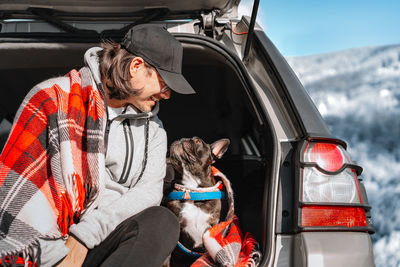 Man with french bulldog dog sitting in car trunk in winter landscape