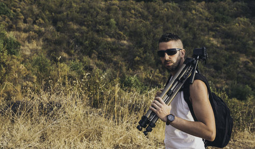 Portrait of mid adult man wearing sunglasses holding tripod while standing on grassy mountain