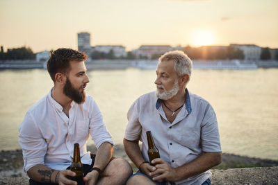 Father and adult son sitting on a wall at the riverside at sunset drinking a beer