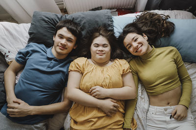 Portrait of happy male and female siblings lying next to sister with down syndrome on bed at home