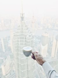 High angle view of hand holding coffee cup against skyscrapers in city