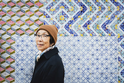 Portrait of senior woman wearing warm clothing standing against colorful tiled wall in city