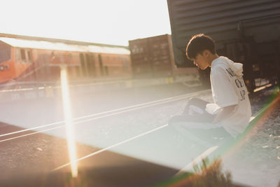 Side view of young man sitting on railroad track against sky during sunny day