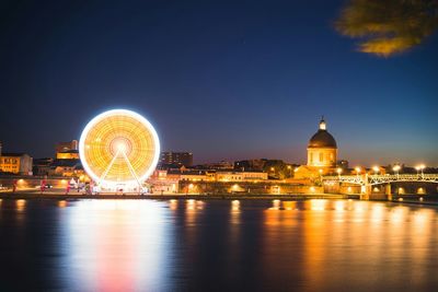 Illuminated ferris wheel and hospital de la grave by river against sky at dusk