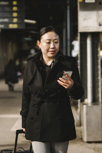 Businesswoman using smart phone while walking with luggage at railroad station