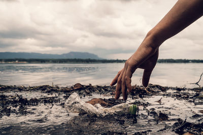 Cropped hand picking plastic bottle from shore at beach against cloudy sky