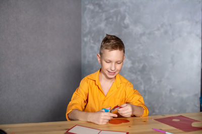 A cute boy is sitting at a table and making valentine's day cards with red hearts