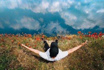 Rear view of man with arms outstretched with poppy flowers on land by lake with clouds reflections