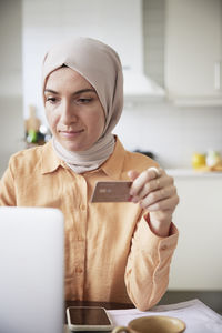 Smiling woman with hijab holding credit card while doing online shopping